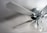 A close-up view of a ceiling fan with four blades and attached light fixtures. The fan and light fixtures are white, with the light fixtures having a floral, frosted glass design. Two pull chains hang from the light fixture. The fan is mounted on a smooth ceiling, casting subtle shadows.
