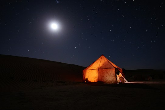 A glowing tent stands illuminated under a clear night sky filled with stars and a bright moon. The tent is situated in what appears to be a desert, with sand dunes visible in the background.