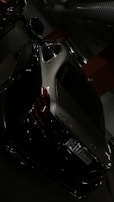 A luxury car, specifically a Maybach, is parked in an indoor garage. The vehicle is dark in color, likely black or dark gray, with sleek curves and reflective surfaces. The environment is dimly lit, with visible concrete and industrial infrastructure, such as large pipes and lighting.