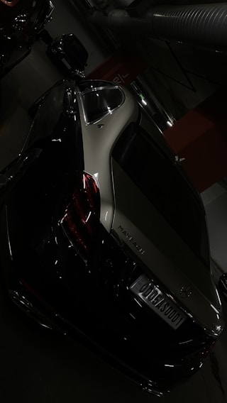 A luxury car, specifically a Maybach, is parked in an indoor garage. The vehicle is dark in color, likely black or dark gray, with sleek curves and reflective surfaces. The environment is dimly lit, with visible concrete and industrial infrastructure, such as large pipes and lighting.