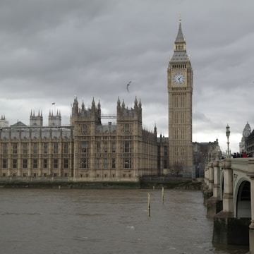 A large, iconic clock tower stands beside a historic parliamentary building, with gothic architectural elements and intricate details. The scene is set against a backdrop of overcast skies, and the structure runs parallel to a wide river. In the distance, a bridge connects the two riverbanks, and a few people can be seen walking on it. A seagull flies overhead, adding a sense of motion to the otherwise still environment.