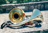 A brass instrument, possibly a sousaphone or tuba, rests on a stone surface outdoors. The shiny metallic surface reflects sunlight, creating bright highlights. The background features a blurred stone wall and greenery.