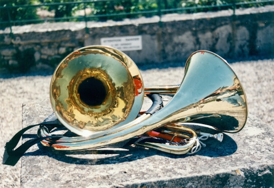 A brass instrument, possibly a sousaphone or tuba, rests on a stone surface outdoors. The shiny metallic surface reflects sunlight, creating bright highlights. The background features a blurred stone wall and greenery.