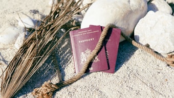 Two dark red passports are placed on a sandy surface surrounded by large white rocks and dry palm leaves. A thick, textured rope winds around the passports, creating a natural and rugged backdrop.