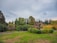 A landscaped garden with neatly cut grass and a variety of lush green trees. Prominent hedges and towering trees form the background, with a sculptural element made of shrub-like formations placed centrally. Bright red flowers add color to the scene.