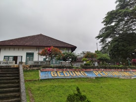 A traditional building with a tiled roof and white walls. There are green doors and windows. In front of the building, there is a garden with vibrant flowers and neatly trimmed hedges. A concrete staircase leads up to the building. The sign in front reads 'GEDUNG PERUNDINGAN'. The sky is overcast, and there are large trees on the right side of the image.