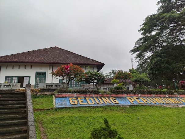 A traditional building with a tiled roof and white walls. There are green doors and windows. In front of the building, there is a garden with vibrant flowers and neatly trimmed hedges. A concrete staircase leads up to the building. The sign in front reads 'GEDUNG PERUNDINGAN'. The sky is overcast, and there are large trees on the right side of the image.