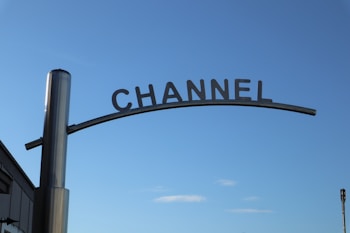 A metallic sign with the word 'CHANNEL' is mounted on a pole against a clear blue sky. The sign curves slightly upward.