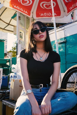 A woman with medium-length black hair is wearing sunglasses, a black top, blue jeans, a necklace, and a watch. She is sitting casually in front of a street vendor cart with red and white umbrellas overhead, marked with the Algida logo. In the background are boxes and bottles, and the side of the cart is painted in teal and white.