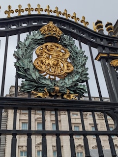 A ornate iron gate featuring a golden monogrammed emblem with a crown on top, surrounded by a detailed wreath. Behind the gate stands a grand building with classical architectural elements such as columns and large windows.