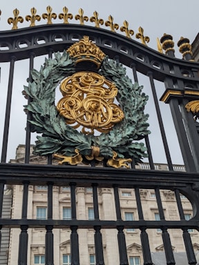 A ornate iron gate featuring a golden monogrammed emblem with a crown on top, surrounded by a detailed wreath. Behind the gate stands a grand building with classical architectural elements such as columns and large windows.