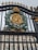 A ornate iron gate featuring a golden monogrammed emblem with a crown on top, surrounded by a detailed wreath. Behind the gate stands a grand building with classical architectural elements such as columns and large windows.