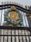 A ornate iron gate featuring a golden monogrammed emblem with a crown on top, surrounded by a detailed wreath. Behind the gate stands a grand building with classical architectural elements such as columns and large windows.