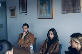 Several people are sitting around a table in a well-lit room. The walls are adorned with abstract paintings. One person is holding their chin thoughtfully, and another has a bottle of water in front of them.
