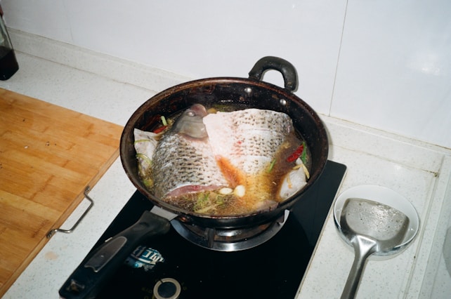 A fish being cooked in a pan on a stovetop, surrounded by herbs and spices. The fish appears to be seasoned with ingredients like garlic and chili peppers. The kitchen counter includes a wooden cutting board and a ladle in a bowl nearby.