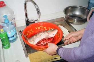 A freshly cleaned fish rests in an ornate red bowl under a running faucet. The sink area includes various cleaning supplies, bottles, and a metal bowl. A person, partially visible, holds the bowl with gloved hands.