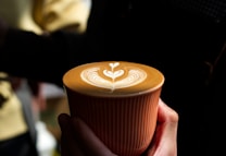A hand holding a cup of coffee adorned with intricate latte art featuring a heart-shaped design. The cup itself is orange with subtle vertical stripes, and the image is captured with a contrasting background that highlights the coffee's foam art.
