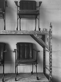 Several rows of dark, vintage-style chairs with a metal frame and padded seats and backs, stacked on industrial metal shelving. The chairs appear slightly worn and are positioned against a plain wall, creating a stark, utilitarian atmosphere.