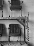 Several rows of dark, vintage-style chairs with a metal frame and padded seats and backs, stacked on industrial metal shelving. The chairs appear slightly worn and are positioned against a plain wall, creating a stark, utilitarian atmosphere.