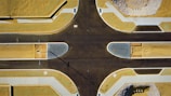 An aerial view of a freshly paved intersection in a developing area. The roads are dark and new, bordered by sandy plots and neatly arranged sidewalks. A long shadow of a streetlight extends across the intersection.