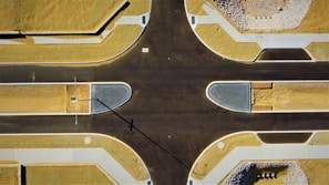 An aerial view of a freshly paved intersection in a developing area. The roads are dark and new, bordered by sandy plots and neatly arranged sidewalks. A long shadow of a streetlight extends across the intersection.