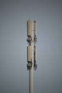 A tall communication tower with multiple antennas and cables stands against a clear sky. The structure is primarily white with dark cables running vertically along it.