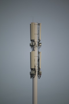 A tall communication tower with multiple antennas and cables stands against a clear sky. The structure is primarily white with dark cables running vertically along it.
