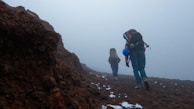 Two hikers are walking up a rocky and foggy trail, carrying large backpacks. Patches of snow are sporadically present on the ground. The terrain appears rugged and challenging.