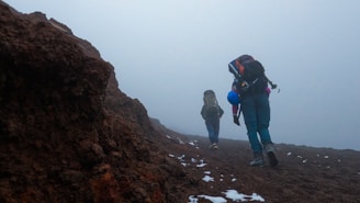 Two hikers are walking up a rocky and foggy trail, carrying large backpacks. Patches of snow are sporadically present on the ground. The terrain appears rugged and challenging.
