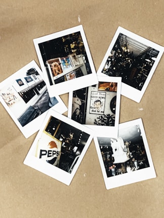 A collection of six Polaroid photographs scattered on a light brown surface. The images include various scenes: a magazine display, a vintage Pepsi sign, an interior with memorabilia, a religious poster, a white statue, and a storefront with awnings and framed pictures. Each photograph captures a moment with nostalgic and eclectic elements.
