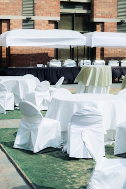 A setup of round tables and chairs covered in white cloth is arranged outdoors on a patch of green artificial turf. Multiple serving stations or buffet tables are positioned in the background under white tents. The setting is adjacent to a brick building.