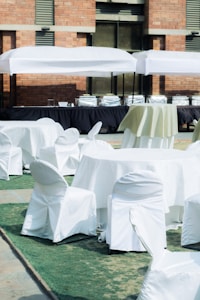 A setup of round tables and chairs covered in white cloth is arranged outdoors on a patch of green artificial turf. Multiple serving stations or buffet tables are positioned in the background under white tents. The setting is adjacent to a brick building.