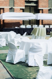A setup of round tables and chairs covered in white cloth is arranged outdoors on a patch of green artificial turf. Multiple serving stations or buffet tables are positioned in the background under white tents. The setting is adjacent to a brick building.