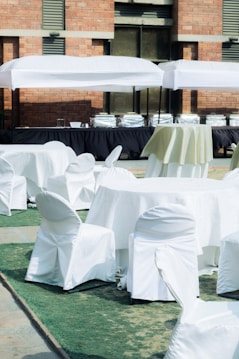 A setup of round tables and chairs covered in white cloth is arranged outdoors on a patch of green artificial turf. Multiple serving stations or buffet tables are positioned in the background under white tents. The setting is adjacent to a brick building.