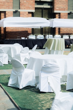 A setup of round tables and chairs covered in white cloth is arranged outdoors on a patch of green artificial turf. Multiple serving stations or buffet tables are positioned in the background under white tents. The setting is adjacent to a brick building.