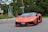 A sleek, orange sports car is navigating a road with lush greenery in the background. The car has a low, aerodynamic design, sharp angles, and a personalized license plate. It is positioned on a paved road with a curb of stone bricks on the side and traffic signs visible.