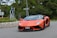 A sleek, orange sports car is navigating a road with lush greenery in the background. The car has a low, aerodynamic design, sharp angles, and a personalized license plate. It is positioned on a paved road with a curb of stone bricks on the side and traffic signs visible.