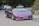 A purple sports car with a customized license plate displaying 'CHILLOUT' is driving on a road. The car is sleek and has a convertible design. Behind the car, there are several cyclists and parked vehicles on a gently curving road. The setting is surrounded by greenery and trees.