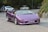 A purple sports car with a customized license plate displaying 'CHILLOUT' is driving on a road. The car is sleek and has a convertible design. Behind the car, there are several cyclists and parked vehicles on a gently curving road. The setting is surrounded by greenery and trees.