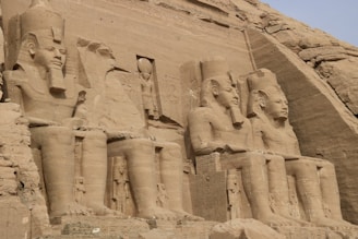 The image displays a series of large stone statues carved into a cliff face, depicting seated pharaohs and adorned with ancient Egyptian hieroglyphs. The detail in the carvings suggests they are part of an archaeological site, likely of historical and cultural significance.