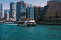 A modern boat travels along a body of turquoise water, surrounded by tall, contemporary skyscrapers and diverse architectural buildings. Palm trees line the waterside promenade, adding a touch of greenery amid the urban setting. The boat appears to be a passenger vessel with people visible on its deck, enjoying the view.