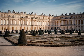 A grand and ornate palace with elaborate architecture stands in the background. The foreground features a formal garden with meticulous hedging and conical topiaries. The sky is clear with a few streaks of clouds, and some people are walking around the garden area.
