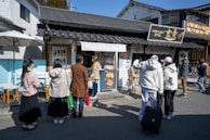 A group of people is standing outside a shop with traditional Japanese architecture, featuring a tiled roof and wooden elements. The shop has a sign with Japanese characters and displays various menu items or advertisements. Some people are looking at the menu, and one person has a suitcase. The setting appears to be on a street with other buildings visible in the background.