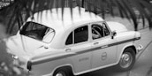 A classic taxi car is parked on the street, partially obscured by foreground foliage. The design of the car is vintage, with rounded edges and a two-tone paint job with the word 'TAXI' visible on the side. The driver seems to be seated inside, positioned at the wheel.