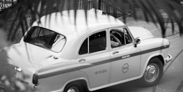 A classic taxi car is parked on the street, partially obscured by foreground foliage. The design of the car is vintage, with rounded edges and a two-tone paint job with the word 'TAXI' visible on the side. The driver seems to be seated inside, positioned at the wheel.