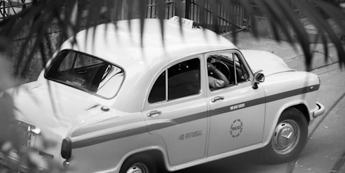 A classic taxi car is parked on the street, partially obscured by foreground foliage. The design of the car is vintage, with rounded edges and a two-tone paint job with the word 'TAXI' visible on the side. The driver seems to be seated inside, positioned at the wheel.
