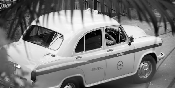 A classic taxi car is parked on the street, partially obscured by foreground foliage. The design of the car is vintage, with rounded edges and a two-tone paint job with the word 'TAXI' visible on the side. The driver seems to be seated inside, positioned at the wheel.