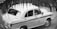 A classic taxi car is parked on the street, partially obscured by foreground foliage. The design of the car is vintage, with rounded edges and a two-tone paint job with the word 'TAXI' visible on the side. The driver seems to be seated inside, positioned at the wheel.