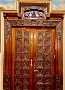 A wooden double door, intricately carved with detailed floral and geometric patterns. The frame features ornate carvings including animal figures and a central decorative panel above the door. The wood appears polished, giving it a rich, dark hue.