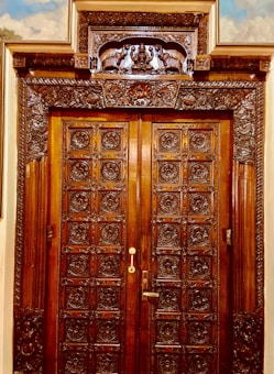 A wooden double door, intricately carved with detailed floral and geometric patterns. The frame features ornate carvings including animal figures and a central decorative panel above the door. The wood appears polished, giving it a rich, dark hue.